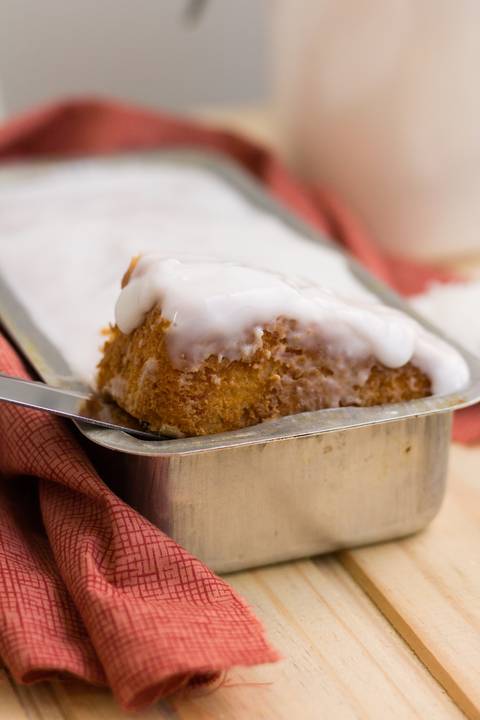 bolo de coco artesanal sobre uma mesa em forma de metal, no bolo vemos uma calda branca e uma fatia de bolo sendo retirada'