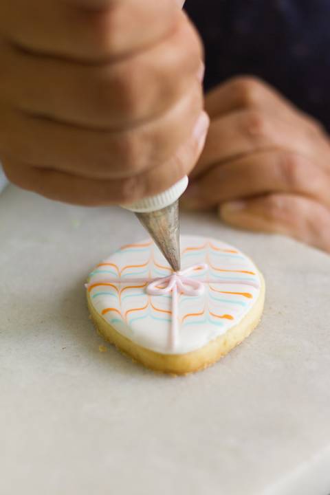 biscoito decorado em forma de ovo de pascoa sendo finalizado. Na imagem vemos o biscoito sob a mesa onde uma mão dá os últimos retoques no biscoito colorido'