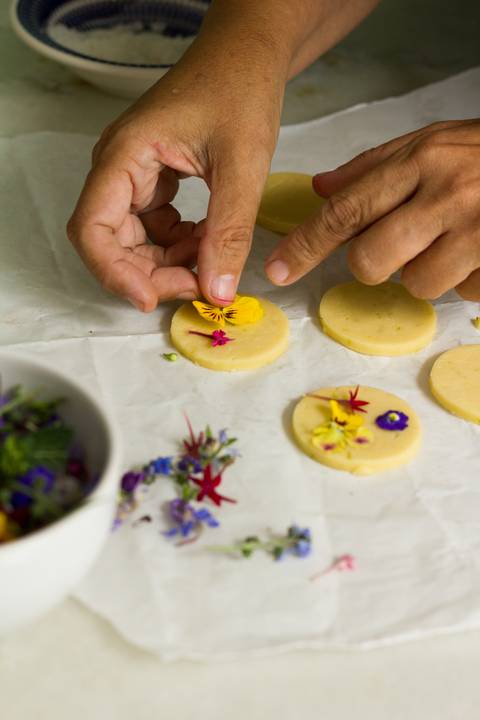 processo de produção dos biscoitos amanteigados com flores comestíveis. Vemos uma mão colocando uma flora amarela sobre massa de biscoito crua '