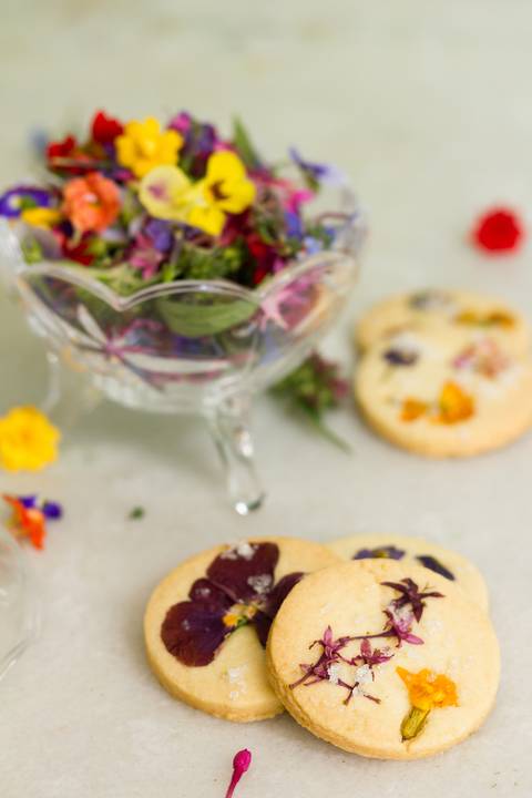 flores comestíveis  espalhadas sobre uma mesa onde também vemos alguns biscoitos para o café da tarde'