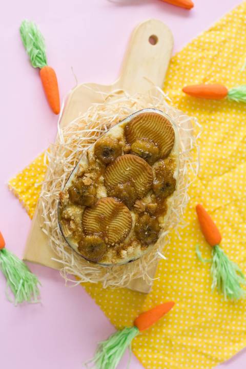 ovo de pascoa artesanal de colher no sabor torta de banana feito pela chef confeitera ludmila colaça, o ovo está sobre um pequeno ninho feito com palha em cima de uma pequena tábua de madeira, vemos também um fundo na cor rosa e diversas cenourinhas '