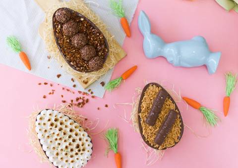 ovos de páscoa sabores diversos espalhados sobre um fundo colorido na cor rosa onde  também vemos um pequeno coelho de porcelana azul e diversas cenourinhas espalhadas por todos os lados'