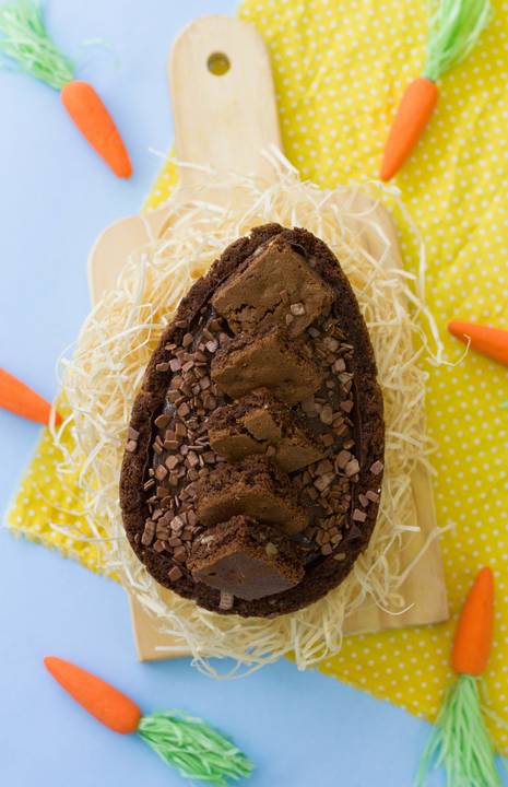 ovo de páscoa de colher sabor brownie colocado dentro de um pequeno ninho feito de palha sobre um fundo colorido de azul e amarelo cercado por diversas cenourinhas'