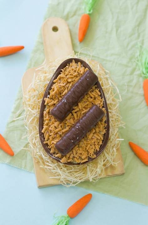 ovo de páscoa artesanal de colher no sabor caramelo salgado sobre um pequeno monte de palha como se fosse um ninho sobre um fundo colorido na cor azul cercado por diversas cenourinhas laranjas'