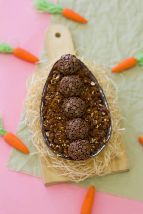 ovo de páscoa artesanal de colher sabor brigadeiro que está colocado sobre um pequeno ninho de palha sobre um fndo rosa com verde cercado de diversas cenourinhas'