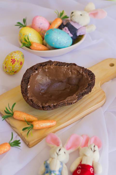 casca de ovo de páscoa sabor brownie colocado sobre uma pequena tábua de madeira em meio a um fundo branco onde vemos alguns adereços de páscoa, cenouras, ovos coloridos'