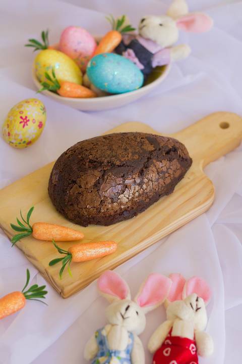 casca de ovo de páscoa sabor brownie colocado sobre uma pequena tábua de madeira em meio a um fundo branco onde vemos alguns adereços de páscoa, cenouras, ovos coloridos'