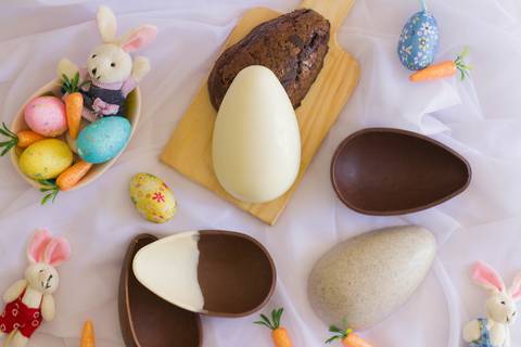 fundo branco onde vemos várias cascas de ovos de páscoa artesanais de diversas cores e sabores rodeados de elementos de páscoa como ovos coloridos, coelhinhos e cenourinhas '