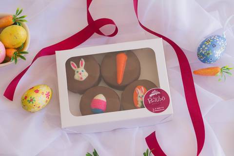 caixa artesanal com quatro alfajores com temática de páscoa, a caixa está sobre um tecido branco tipo voal e rodeada por uma fita de cor marsala e elementos de páscoa como ovinhos coloridos e cenourinhas'