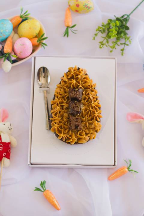 ovo de páscoa de colher artesanal centralizado na imagem, o ovo está dentro de uma caixa de papel branca ao lado vemos uma pequena colher, essa caixa centralizada está rodeada de elementos de páscoa como coelhinhos de pelúcia e cenourinhas'