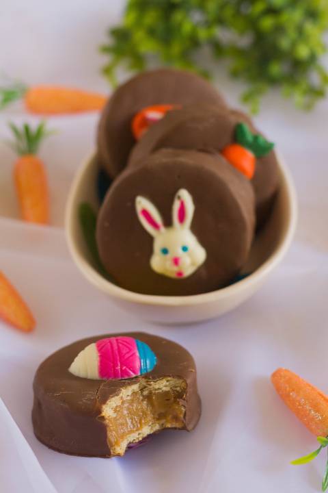 alfajor artesanal com temática de páscoa dentro de um pequeno pote de louça no formato de ovo de páscoa, mais a frente vemos um alfajor mordido rodeados de elementos de páscoa como cenourinhas e ovinhos de páscoa'