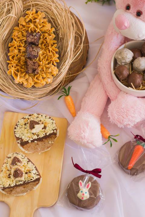 cenário de páscoa onde vemos alguns doces de páscoa como ovos de colher e alfajores, na imagem no canto superior direito vemos um grande coelho de páscoa rosa segurando um pote com algumas trufas de palha italiana'