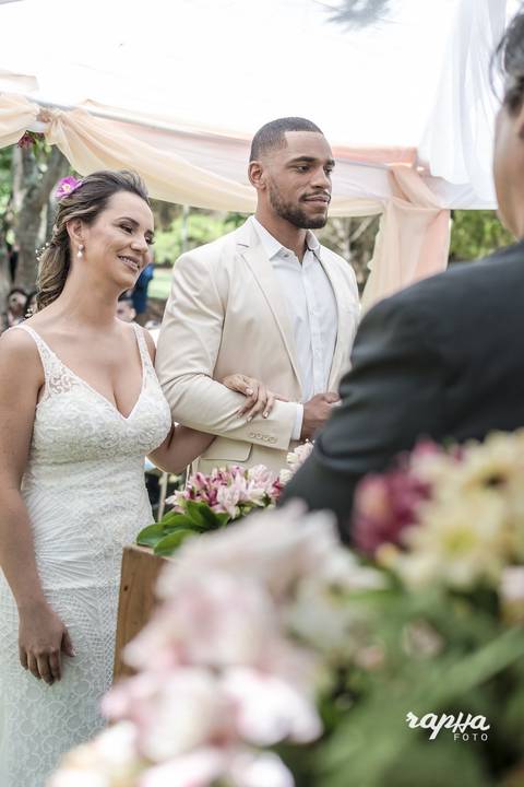 Noivos no altar, a noiva com vestido todo branco e o noivo com traje também claro, fotografado pelo Rapha Drummond de Belo Horizonte.'
