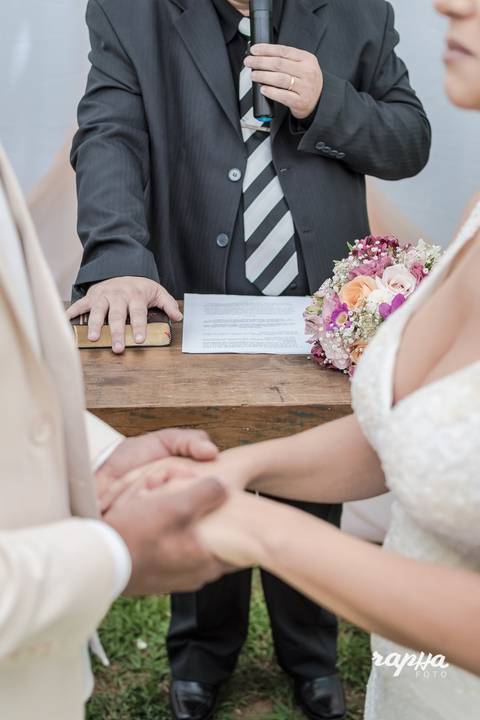 Fotógrafo de casamentos em BH, Rapha Drummond fotografa noivos de mão dadas em segundo plano enquanto o foco está na biblía na mesa com a mão do pastor pousada em cima.'