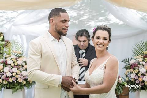 Fotógrafo de casamentos registra o momento onde o noivo olha apaixonadamente para sua amada e a noiva por sua vez olha encantada para daminha que está trazendo as alianças;'