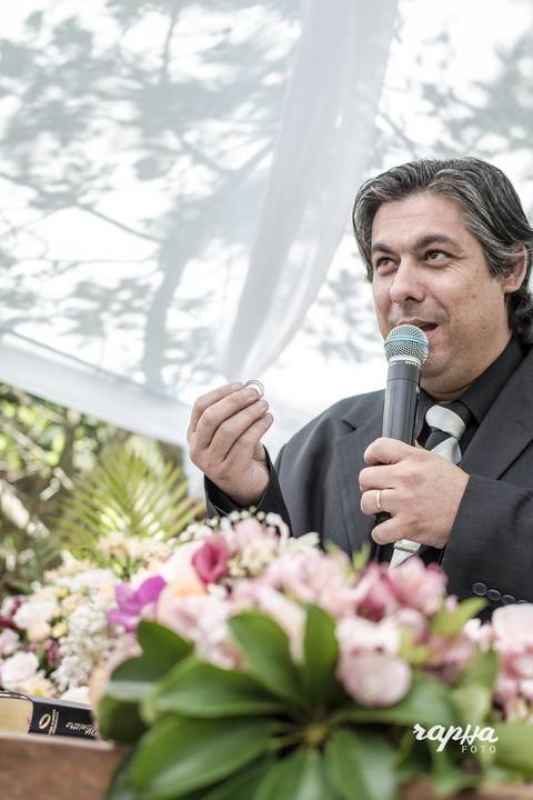 Fotóografo de Casamentos registra pastor erguendo sua mão direita com o par de alianças. Flores e cores em destaque.'