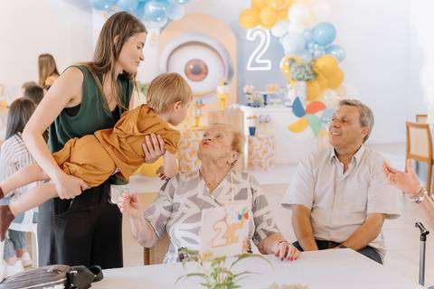 Léo no colo da bisavó com familiares ao redor, durante a festa de aniversário em Mogi'