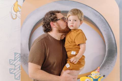 Pai dando um beijo no Léo ao lado da decoração do aniversário infantil'