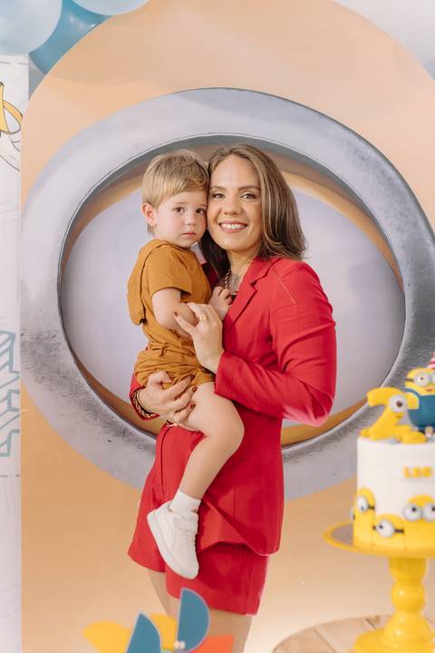 Léo no colo da mãe, com fundo decorado com Minions no salão de festas'