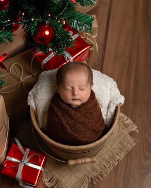 Ensaio de Recém-nascido. Bebê dormindo em baldinho em ensaio Newborn no Estúdio Amoradia Foto. Ao redor tem luzes e presentes natalinos.'