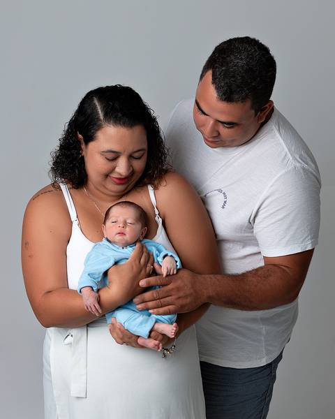 Ensaio Newborn Natal. Foto de recém-nascido sendo segurado pela mãe, o pai olha para os dois.'