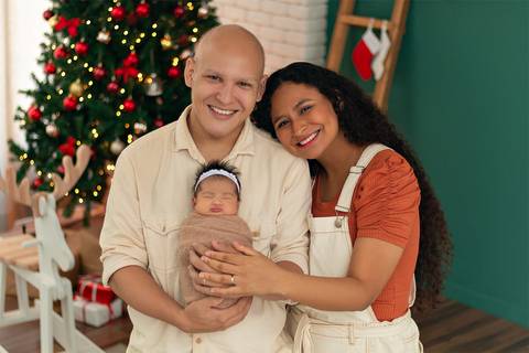 Bebê recém-nascida com pai e mãe em fotos em estúdio com cenário natalino.'