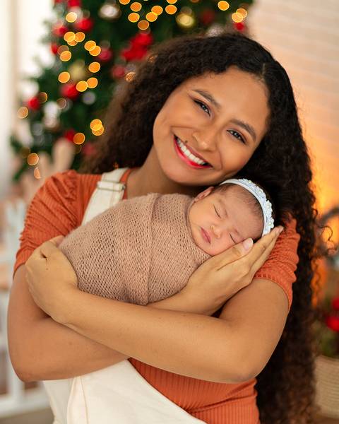 Bebê recém-nascida com mãe em fotos em estúdio com cenário natalino.'