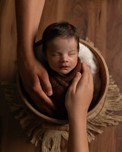 O bebê recém-nascido está dormindo enroladinho no wrap em seu ensaio de primeiros dias. As mãos dos pais estão em volta do recém-nascido.'