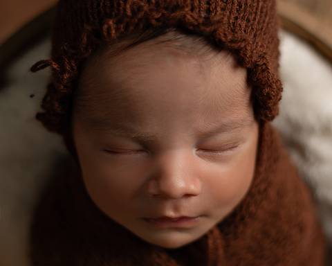 O bebê recém-nascido está dormindo enroladinho no wrap usando uma toquinha em seu ensaio de primeiros dias. '