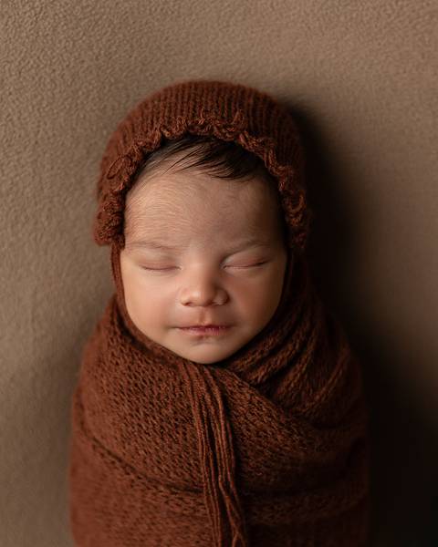 O bebê recém-nascido está dormindo enroladinho no wrap usando uma toquinha em seu ensaio de primeiros dias. '