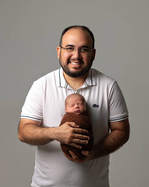 Ensaio Newborn em estúdio. Bebê recém-nascido enrolado no wrap nos braços do pai.'