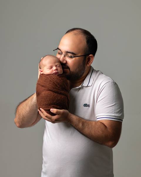 Ensaio Newborn em estúdio. Bebê recém-nascido enrolado no wrap nos braços do pai.'