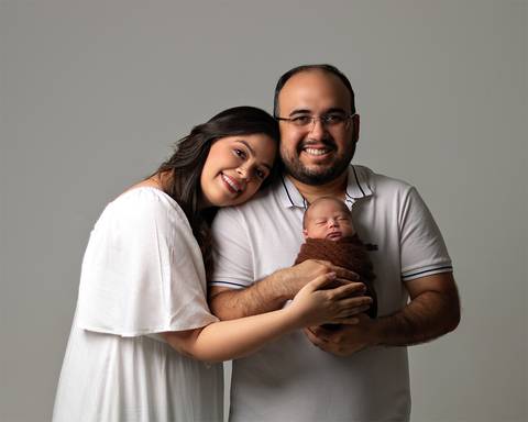 Ensaio Newborn em estúdio. Bebê recém-nascido enrolado no wrap nos braços dos pais.'