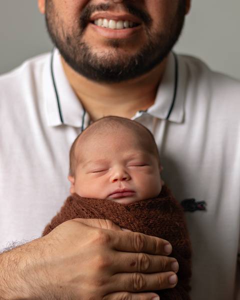 Ensaio Newborn em estúdio. Bebê recém-nascido enrolado no wrap nos braços do pai.'