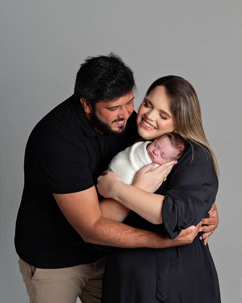 Ensaio Newborn. Bebê enrolado no wrap sendo segurado pela mãe e o pai.'