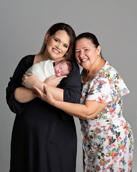 Ensaio Newborn. Bebê enrolado no wrap sendo segurado pela mãe e a avó.'