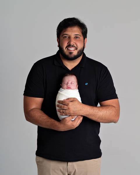 Ensaio Newborn. Bebê enrolado no wrap sendo segurado pelo pai.'