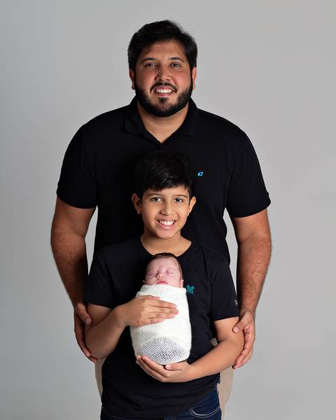 Ensaio Newborn. Bebê enrolado no wrap sendo segurado pelo pai e o irmão.'