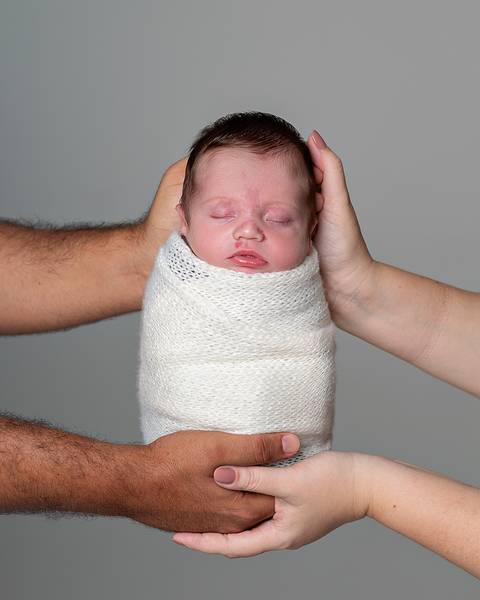 Ensaio Newborn. Bebê enrolado no wrap sendo segurado pelas mãos dos pais.'