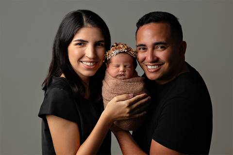 Mãe e pai segurando filha recém-nascida enrolada no wrap em ensaio Newborn em estúdio.'