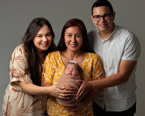 Bebê dormindo nos braços da avó em ensaio newborn. Os pais estão ao lado.'