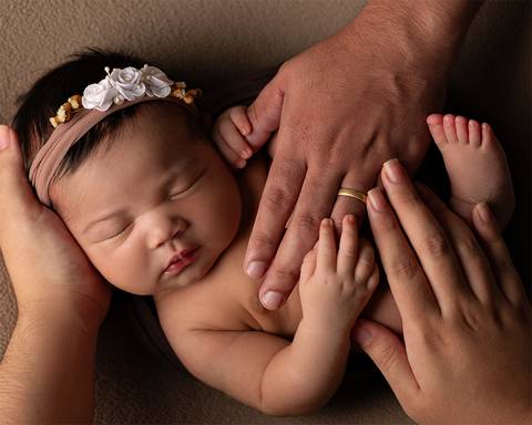 Bebê dormindo em ensaio newborn, as mãos dos pais estão sobre ela.'