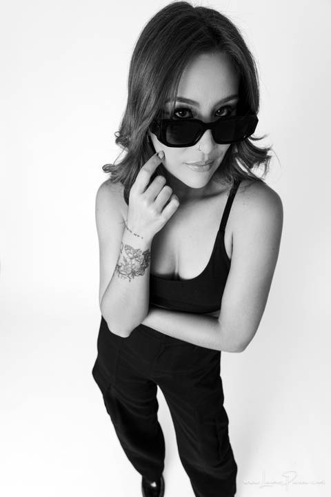  mulher em pé olhado por cima do oculos e com mão no rosto, usando top e calça cargo preta, no fundo branco para retrato feminino em estudio fotografico para fotos corporativas em natal'