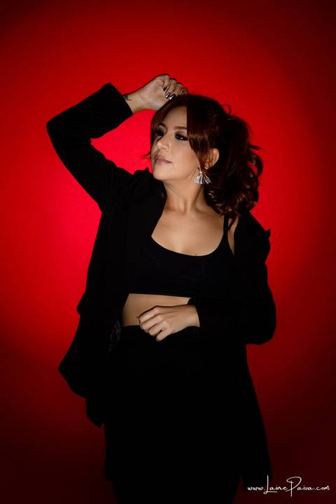  mulher encostada na parede em pé com mão na barriga e uma para cima usando blaze, top e calça cargo preta, no fundo vermelho para retrato feminino em estudio fotografico para fotos corporativas em natal'