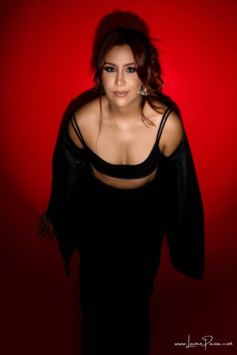  mulher encostada na parede em pé com mãos para trás usando blaze, top e calça cargo preta, no fundo vermelho para retrato feminino em estudio fotografico para fotos corporativas em natal'