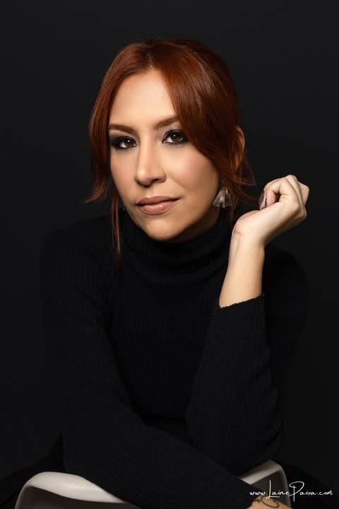  mulher  sentada em cadeira com um braço apoiado e outro proximo ao rosto olhando para o lado usando blusa gola alta e calça cargo preta, no fundo cinza para retrato feminino em estudio fotografico para fotos corporativas em natal'