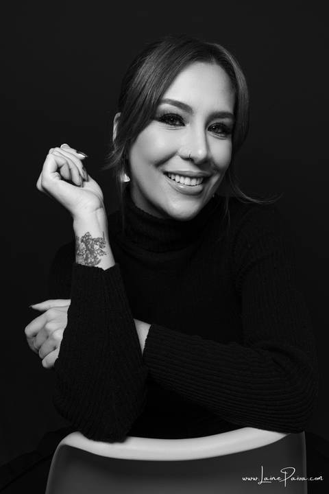  mulher sorrindo sentada em cadeira com um braço apoiado e outro proximo ao rosto olhando para o lado usando blusa gola alta e calça cargo preta, no fundo cinza para retrato feminino em estudio fotografico para fotos corporativas em natal'