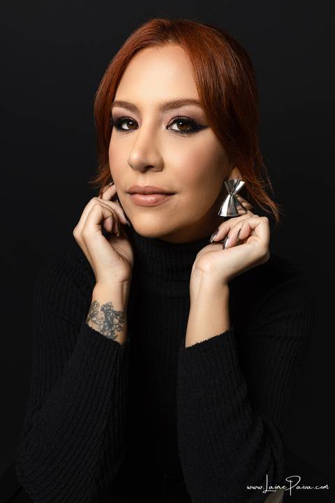  mulher com mãos tocando em brincos, olhando para o lado usando blusa gola alta e calça cargo preta, no fundo cinza para retrato feminino em estudio fotografico para fotos corporativas em natal'