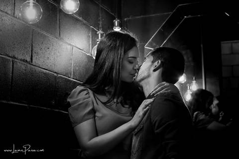 Mulher sentada e homem entre suas pernas abraçando-a e quase se beijando com luzes sob a cabeça deles em banheiro escuro do loft de macamirim'