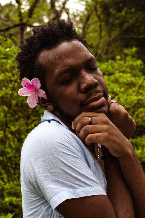 Hugo em frente a parede vermelha de tijolos no parque do carmo, na capital de são paulo. Em frente aos arbustos, com uma flor na cabeça, enquanto fecha os olhos, enquadramento mais em seu rosto.'